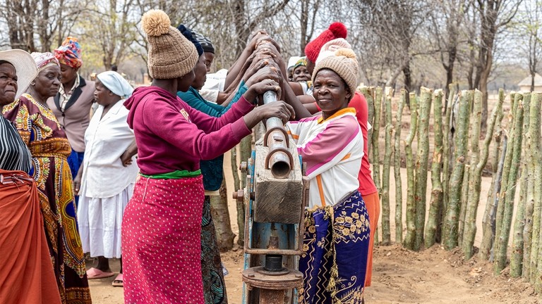 A group of women in bright coloured clothing pumping water at a water pump in Zimbabwe