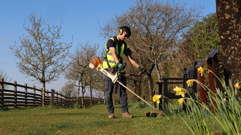 An image of a man using a strimmer to cut grass around a bench on a grass walkway.