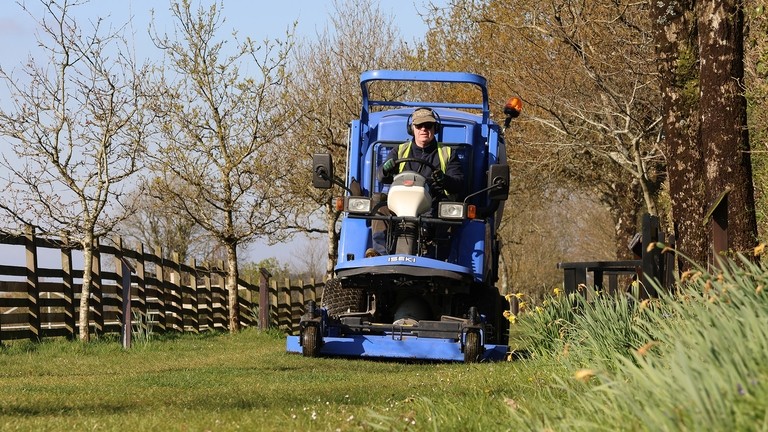 An image of a man sat on a blue lawn mower machine, driving down a grass walkway.