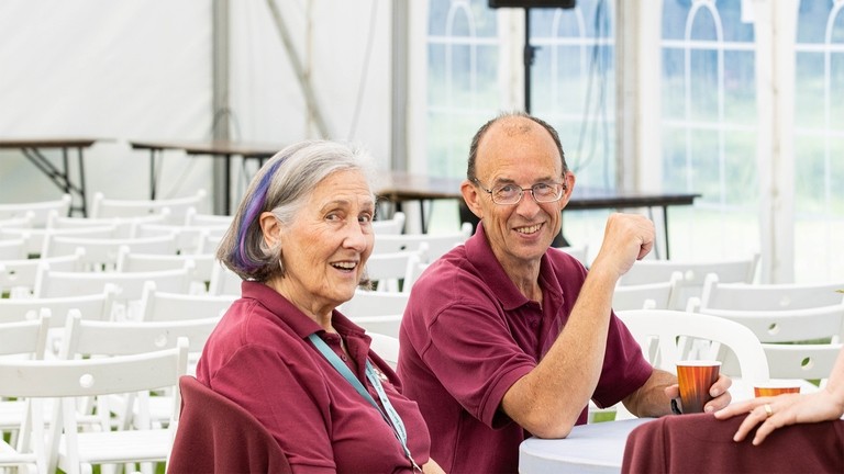 An image of a women and man wearing burgundy polo shirts, sat looking toward the camera.