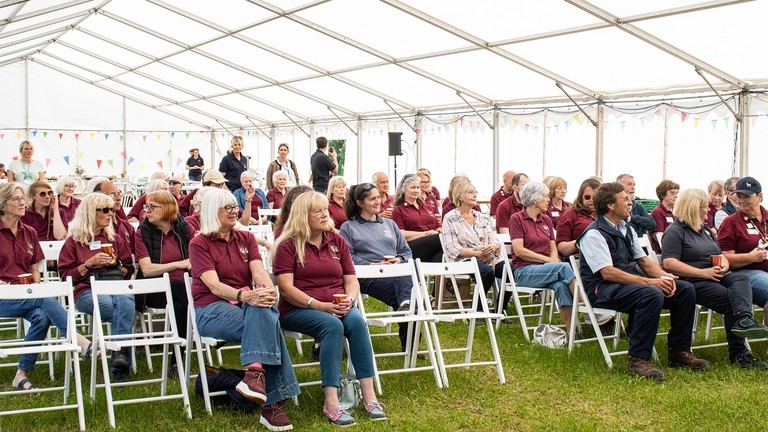 An image of a group of people wearing burgundy polo shirts, sat on rows of white chairs under a white marquee.