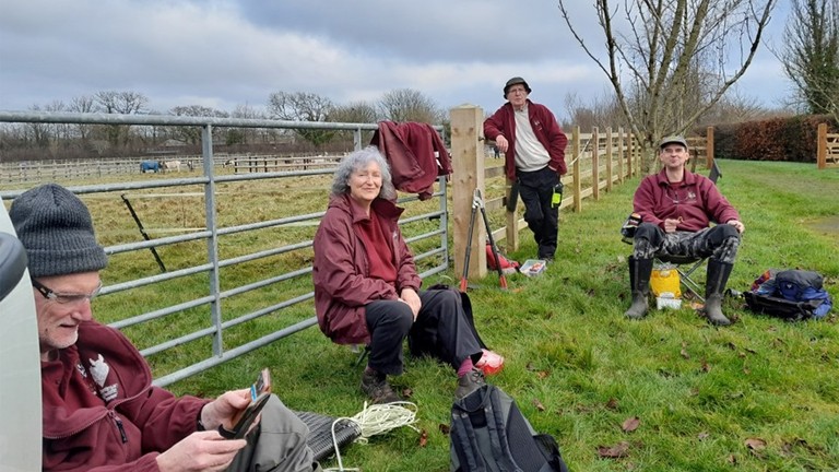 An image of a group of people wearing burgundy fleeces while sat resting next to a fence.