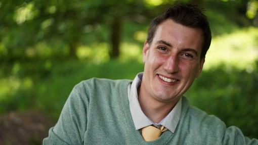 A headshot image of a brown haired man wearing a green jumper and yellow tie.