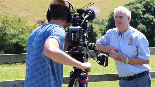 A man holding a camera filming an older man leaning against a fence