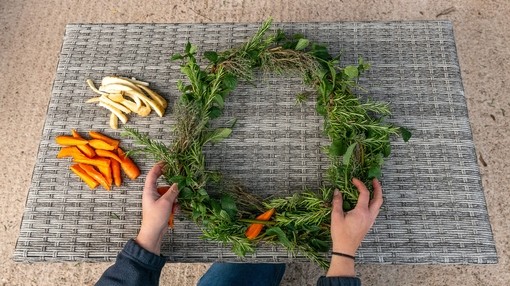 A pair of hands holding a partially finished wreath with a pile of carrots and parsnips next to it