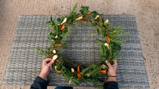 A pair of hands holding a finished wreath featuring multiple green leaves, carrots and parsnips