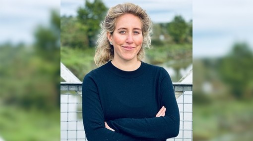 A blonde women wearing a long black t-shirt with her arms crossed smiling at the camera