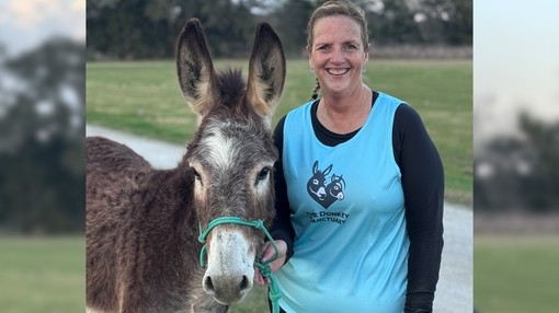 A women wearing a blue running top with a donkey logo stood next to a large grey donkey with a white nose.