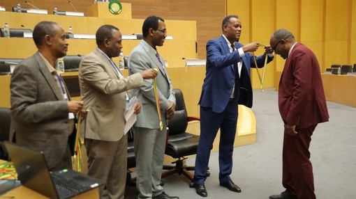A man in a blue suit presenting a medal to a man in a red suit as three other men watch and smile.