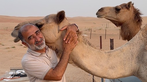 An image of a man hugging the neck of a camel as it rest its head on the mans shoulder while another camel stands behind them.