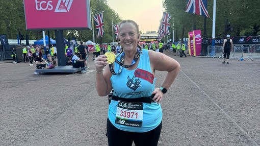 An image of a women wearing a blue runner top holding a gold medal as she poses in front of a marathon finish line.