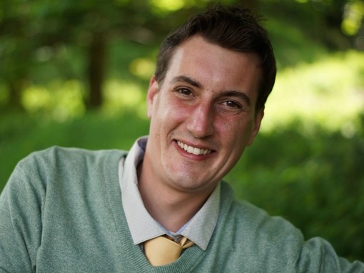A headshot image of a brown haired man wearing a green jumper and yellow tie.
