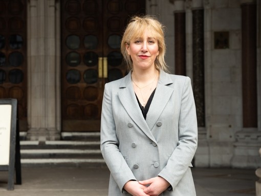 A women dressed in a smart grey blazer stood outside the entrance of a concrete building with two wooden doors.
