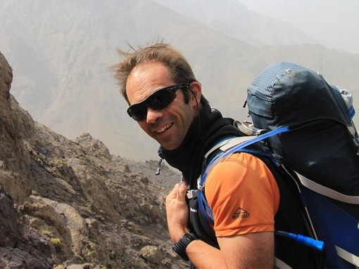 An image of man wearing an orange top and rucksack looking toward the camera as he poses on a mountain side whilst trekking.