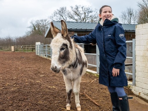 An image of a girl wearing a coat and wellies stood with a white and grey skewbald donkey in an outside woodchip paddock.